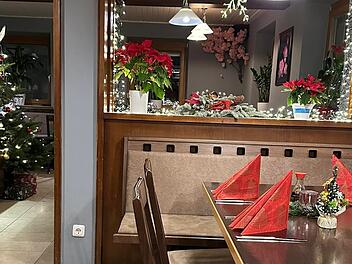 Ein gem&uuml;tlich dekorierter Raum mit Weihnachtsthema. Ein gedeckter Tisch mit roten Servietten, Weihnachtsschmuck und einem Weihnachtsbaum mit Geschenken im Hintergrund.