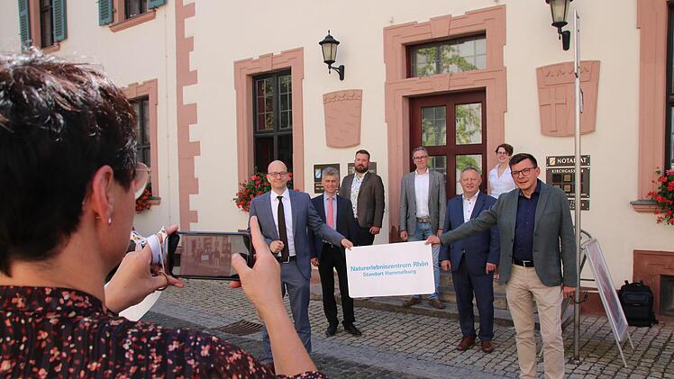 Gruppenbild von der offiziellen Eröffnung am Eingang zum Roten Schloss in Hammelburg: Der Bad Kissinger OB Dirk Vogel (links) und der Hammelburger Bürgermeister Armin Warmuth (rechts) halten das neue NEZ-Schild. Dahinter Abteilungsdirektor Bertram Eidel und Landrat Thomas Bold, auf der Treppe die Mitarbeiter Felix Papsch, Dr. Joachim Schneider und Lisa Graskamp. Foto: Ralf Ruppert