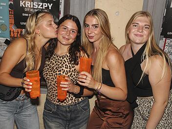 Vier junge Frauen feiern und halten orangefarbene Becher. Sie sind fr&ouml;hlich und ausgelassen. Im Hintergrund sind Plakate zu sehen.