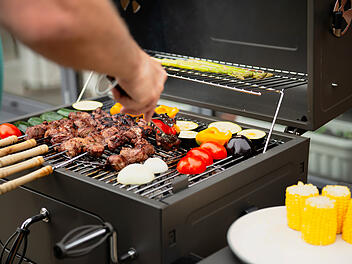 Auf einem Elektrogrill liegen mehrere Grillspie&szlig;e und Grillgem&uuml;se. Daneben steht ein Teller mit Essmais.