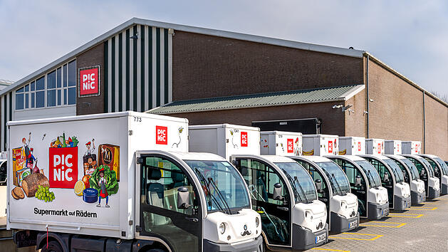 Eine Reihe von Lieferwagen mit der Aufschrift "PIC NIC" steht vor einem Gebäude. Die Fahrzeuge sind für einen Online-Supermarkt, der als "Supermarkt auf Rädern" wirbt.