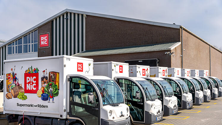 Eine Reihe von Lieferwagen mit der Aufschrift "PIC NIC" steht vor einem Gebäude. Die Fahrzeuge sind für einen Online-Supermarkt, der als "Supermarkt auf Rädern" wirbt.