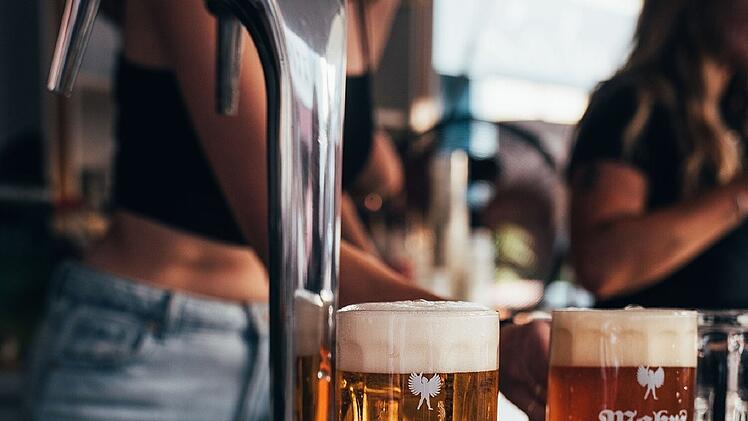 Zwei gefüllte Bierkrüge mit dem Logo "Mahrs Bräu Bamberg" stehen auf einer Theke in einer Bar oder Brauerei. Im Hintergrund sind zwei Personen sichtbar.