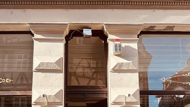 Ein Caf&eacute;-Eingang mit zwei schwarzen Tafeln, verziert mit Schrift. Innen ist ein stilvolles Ambiente und ein Kronleuchter erkennbar. Das Caf&eacute; signalisiert den Beginn eines neuen Kapitels.