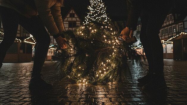 Erstelle ein spontanes Smartphone-Foto (16:9) vom n&auml;chtlichen Marienplatz in Kronach, Deutschland. Die Beleuchtung ist sehr sp&auml;rlich. Im Vordergrund: Zwei junge Personen von hinten &ndash; eine junge Frau und ein junger Mann &ndash; in l&auml;ssigen Winterjacken und M&uuml;tzen. Jeder von ihnen tr&auml;gt einen festlich geschm&uuml;ckten Weihnachtsbaum auf der Schulter. Die B&auml;ume sind sichtlich schwer und werden mit beiden H&auml;nden getragen. Die Szene spielt sich zwischen den dunklen Buden des Weihnachtsmarktes ab, die wenigen Lichter im Hintergrund verschwimmen. Ansonsten befinden sich keine Menschn im Bild. Typisch deutsche Innenstadtarchitektur rahmt den Platz, Kopfsteinpflaster reflektiert die bunten Lichter, es befinden sich keine Menschen auf dem Platz. Die Aufnahme ist stark k&ouml;rnig, mit deutlichem Rauschen und leichter Bewegungsunsch&auml;rfe, als w&auml;re sie hastig und unbemerkt aus der H&uuml;fte geschossen worden. Authentischer, ungeschliffener Look, dokumentarischer Stil, aufgenommen mit einer 28-mm-Linse aus mittlerer Entfernung, geradeaus. Nachtstimmung, sehr schwaches, st&auml;dtisches Licht, die Lichterketten der Christb&auml;ume sorgen f&uuml;r kleine Lichtinseln im Bild.&nbsp;