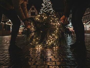 Erstelle ein spontanes Smartphone-Foto (16:9) vom n&auml;chtlichen Marienplatz in Kronach, Deutschland. Die Beleuchtung ist sehr sp&auml;rlich. Im Vordergrund: Zwei junge Personen von hinten &ndash; eine junge Frau und ein junger Mann &ndash; in l&auml;ssigen Winterjacken und M&uuml;tzen. Jeder von ihnen tr&auml;gt einen festlich geschm&uuml;ckten Weihnachtsbaum auf der Schulter. Die B&auml;ume sind sichtlich schwer und werden mit beiden H&auml;nden getragen. Die Szene spielt sich zwischen den dunklen Buden des Weihnachtsmarktes ab, die wenigen Lichter im Hintergrund verschwimmen. Ansonsten befinden sich keine Menschn im Bild. Typisch deutsche Innenstadtarchitektur rahmt den Platz, Kopfsteinpflaster reflektiert die bunten Lichter, es befinden sich keine Menschen auf dem Platz. Die Aufnahme ist stark k&ouml;rnig, mit deutlichem Rauschen und leichter Bewegungsunsch&auml;rfe, als w&auml;re sie hastig und unbemerkt aus der H&uuml;fte geschossen worden. Authentischer, ungeschliffener Look, dokumentarischer Stil, aufgenommen mit einer 28-mm-Linse aus mittlerer Entfernung, geradeaus. Nachtstimmung, sehr schwaches, st&auml;dtisches Licht, die Lichterketten der Christb&auml;ume sorgen f&uuml;r kleine Lichtinseln im Bild.&nbsp;