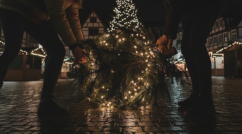 Erstelle ein spontanes Smartphone-Foto (16:9) vom n&auml;chtlichen Marienplatz in Kronach, Deutschland. Die Beleuchtung ist sehr sp&auml;rlich. Im Vordergrund: Zwei junge Personen von hinten &ndash; eine junge Frau und ein junger Mann &ndash; in l&auml;ssigen Winterjacken und M&uuml;tzen. Jeder von ihnen tr&auml;gt einen festlich geschm&uuml;ckten Weihnachtsbaum auf der Schulter. Die B&auml;ume sind sichtlich schwer und werden mit beiden H&auml;nden getragen. Die Szene spielt sich zwischen den dunklen Buden des Weihnachtsmarktes ab, die wenigen Lichter im Hintergrund verschwimmen. Ansonsten befinden sich keine Menschn im Bild. Typisch deutsche Innenstadtarchitektur rahmt den Platz, Kopfsteinpflaster reflektiert die bunten Lichter, es befinden sich keine Menschen auf dem Platz. Die Aufnahme ist stark k&ouml;rnig, mit deutlichem Rauschen und leichter Bewegungsunsch&auml;rfe, als w&auml;re sie hastig und unbemerkt aus der H&uuml;fte geschossen worden. Authentischer, ungeschliffener Look, dokumentarischer Stil, aufgenommen mit einer 28-mm-Linse aus mittlerer Entfernung, geradeaus. Nachtstimmung, sehr schwaches, st&auml;dtisches Licht, die Lichterketten der Christb&auml;ume sorgen f&uuml;r kleine Lichtinseln im Bild.&nbsp;