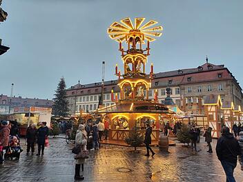 Ein festlich beleuchteter Weihnachtsmarkt mit einer großen Weihnachtspyramide im Mittelpunkt. Menschen schlendern herum, und ein beleuchteter Weihnachtsbaum steht im Hintergrund.