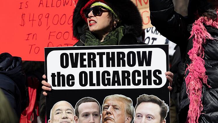 Es rumort in den USA.  Diese Demonstrantin hält ein Schild mit der Aufschrift "Stürzt die Oligarchen" in die Höhe. Das Plakat zeigt die Köpfe von Jeff Bezos (Amazon), Mark Zuckerberg (Facebook, Instagram, WhatsApp), Präsident Trump und Elon Musk (Tesla, X, Space X).