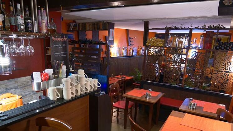 Gemütliches Restaurant mit Holztischen, roten Stühlen, und orangenen Tischsets. Barbereich mit Gläsern und Krügen. Dekorative Trennwand im Hintergrund. Warme, einladende Atmosphäre.