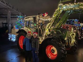 Weihnachtlich dekorierte Traktoren, mit Lichtern geschmückt, stehen in Hallstadt. Zwei Personen in Weihnachtsmützen posieren vor einem Traktor. Ein Baum mit Geschenk ist ebenfalls zu sehen.