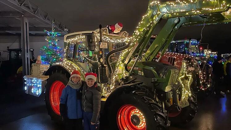 Weihnachtlich dekorierte Traktoren, mit Lichtern geschmückt, stehen in Hallstadt. Zwei Personen in Weihnachtsmützen posieren vor einem Traktor. Ein Baum mit Geschenk ist ebenfalls zu sehen.