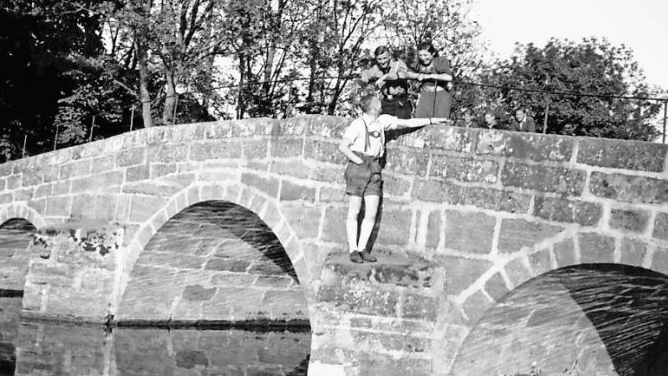 Über die Alster in Memmelsdorf führte früher eine Steinbrücke mit drei Bögen. Sie ist längst von der Bildfläche verschwunden. Das Bild aus dem Jahr 1942 zeigt auf der Brücke im Gespräch mit jungen Männern Agnes Zeuner, die Tochter des einstigen Forstbüttners.  Archivfoto: Arnold Deininger