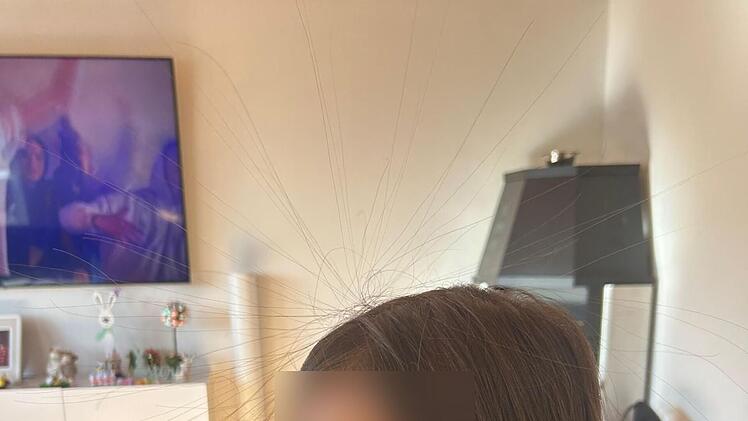 Ein Kind sitzt auf einem Sofa, im Hintergrund ein Fernseher und Deko. Es gibt Kartons neben dem Kind. Haare stehen durch statische Elektrizität ab.