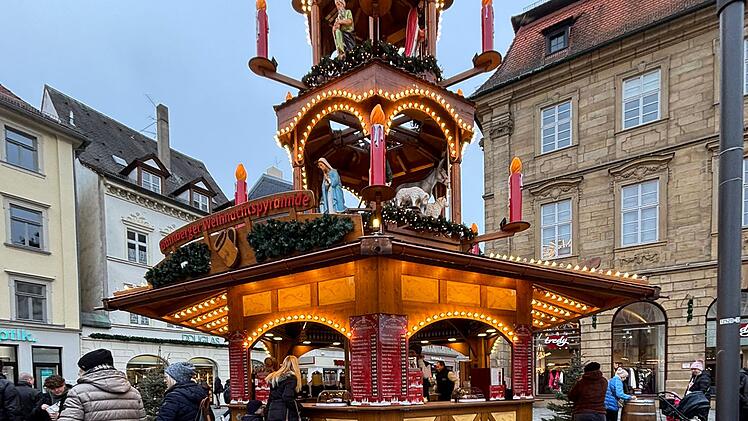 Ein Weihnachtsmarktstand mit leuchtendem Weihnachtsbaum, umgeben von Menschen, die sich versammeln. Pflastersteine und historische Gebäude im Hintergrund.