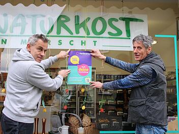 Arno Reuscher (links) und Armin Berninger hängen ein Tante-Enso-Plakat am Naturkostladen auf. Die Ladenbesitzer unterstützen die Neuansiedlung eines Mini-Supermarktes in vollem Umfang. Foto: Thomas Malz