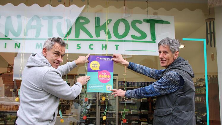 Arno Reuscher (links) und Armin Berninger hängen ein Tante-Enso-Plakat am Naturkostladen auf. Die Ladenbesitzer unterstützen die Neuansiedlung eines Mini-Supermarktes in vollem Umfang. Foto: Thomas Malz