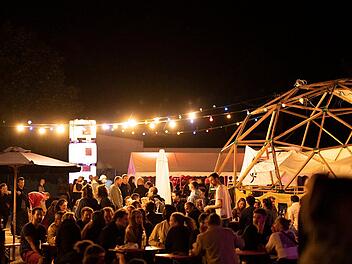 Beim Bamberger Kulturfestival "Kontakt" sitzen Menschen unter bunten Lichtern in geselliger Atmosphäre. Eine Holzkonstruktion und Tische schaffen ein lebendiges Ambiente.