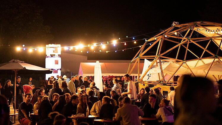Beim Bamberger Kulturfestival "Kontakt" sitzen Menschen unter bunten Lichtern in geselliger Atmosphäre. Eine Holzkonstruktion und Tische schaffen ein lebendiges Ambiente.