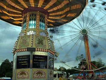 Ein leuchtendes Karussell dreht sich vor einem Riesenrad. Die Lichter des Volksfests erhellen den bew&ouml;lkten Himmel. Atmosph&auml;re von Nostalgie und Freude.