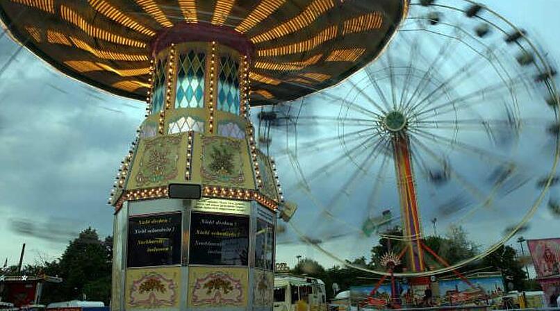 Ein leuchtendes Karussell dreht sich vor einem Riesenrad. Die Lichter des Volksfests erhellen den bew&ouml;lkten Himmel. Atmosph&auml;re von Nostalgie und Freude.