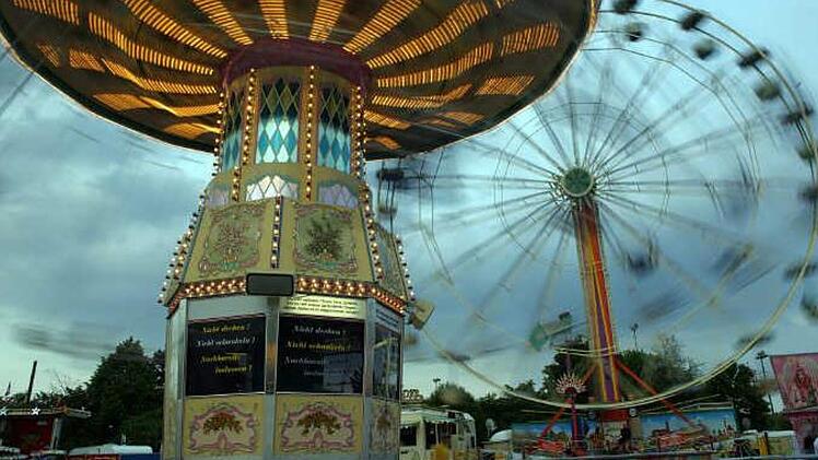 Ein leuchtendes Karussell dreht sich vor einem Riesenrad. Die Lichter des Volksfests erhellen den bewölkten Himmel. Atmosphäre von Nostalgie und Freude.