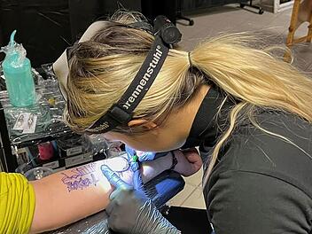 Eine Person tätowiert einen Arm in einem Studio. Sie trägt eine Stirnlampe und ist konzentriert bei der Arbeit. Im Hintergrund sind Tattoo-Utensilien sichtbar.
