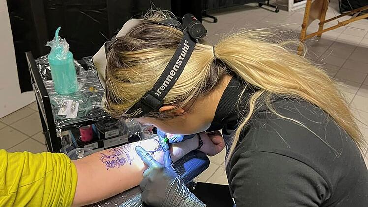 Eine Person tätowiert einen Arm in einem Studio. Sie trägt eine Stirnlampe und ist konzentriert bei der Arbeit. Im Hintergrund sind Tattoo-Utensilien sichtbar.