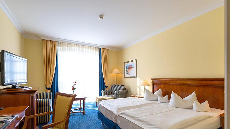 Helles Hotelzimmer mit Doppelbett, blauen Vorhängen und Teppich. Es gibt einen Holzschreibtisch, Sessel und eine Kommode mit TV. Elegantes Ambiente, passend für 4-Sterne-Bereich.