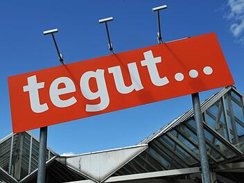 Ein rotes Tegut-Schild vor einem blauen Himmel und einem modernen Glasbau. Die Supermarktkette plant, Filialen zu schließen und zu verkaufen.