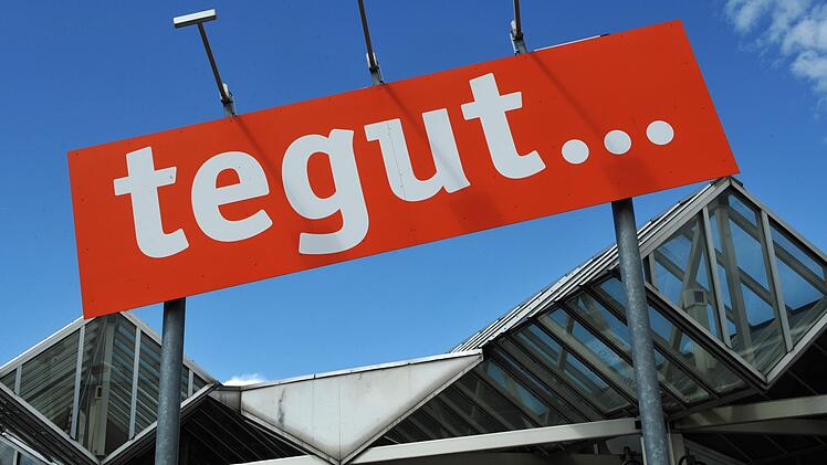 Ein rotes Tegut-Schild vor einem blauen Himmel und einem modernen Glasbau. Die Supermarktkette plant, Filialen zu schließen und zu verkaufen.