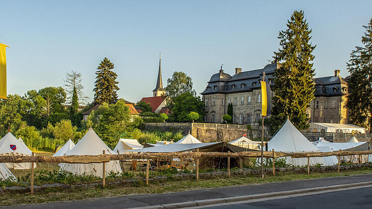 Ein mittelalterliches Lager mit weißen Zelten, Fahnen und einer Burg im Hintergrund. Das Bild zeigt das Pilgerlager in Burgwindheim, wo "Gewandete" im Juni 2025 ein historisches Erlebnis bieten.