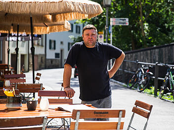 Ein Mann steht drau&szlig;en vor einem Restaurant mit leeren Tischen und Schirmen. Im Hintergrund ist eine gesperrte Br&uuml;cke. Fahrr&auml;der lehnen am Gel&auml;nder.