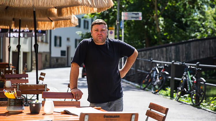 Ein Mann steht drau&szlig;en vor einem Restaurant mit leeren Tischen und Schirmen. Im Hintergrund ist eine gesperrte Br&uuml;cke. Fahrr&auml;der lehnen am Gel&auml;nder.