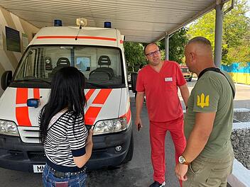 Drei Personen stehen bei einem Rettungswagen. Eine trägt rote Kleidung, die andere hat ein T-Shirt mit weißem Symbol. Hintergrund: Bäume und ein farbiger Zaun. Thema: Unterstützung der Ukraine.