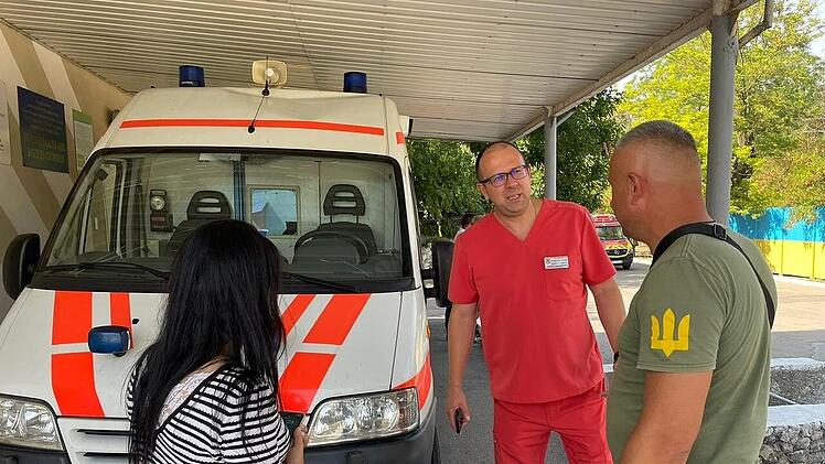 Drei Personen stehen bei einem Rettungswagen. Eine trägt rote Kleidung, die andere hat ein T-Shirt mit weißem Symbol. Hintergrund: Bäume und ein farbiger Zaun. Thema: Unterstützung der Ukraine.