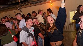 Eine Gruppe fröhlicher junger Menschen auf einem Fest. Eine Frau hebt die Hand und lacht. Getränke stehen auf den Tischen. Stimmung ist ausgelassen und gesellig.