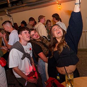 Eine Gruppe fröhlicher junger Menschen auf einem Fest. Eine Frau hebt die Hand und lacht. Getränke stehen auf den Tischen. Stimmung ist ausgelassen und gesellig.