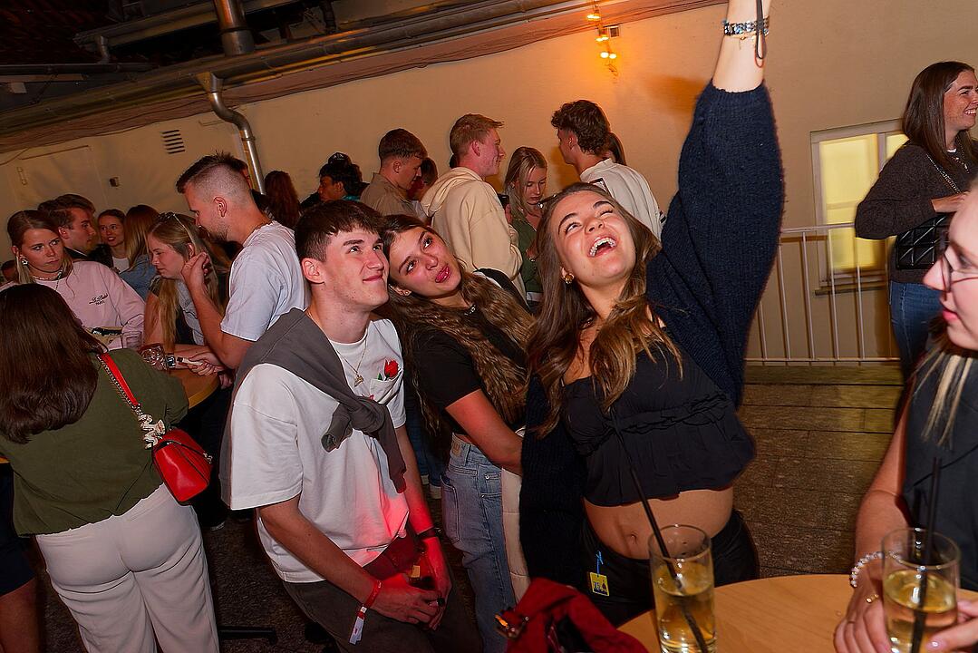 Eine Gruppe fröhlicher junger Menschen auf einem Fest. Eine Frau hebt die Hand und lacht. Getränke stehen auf den Tischen. Stimmung ist ausgelassen und gesellig.