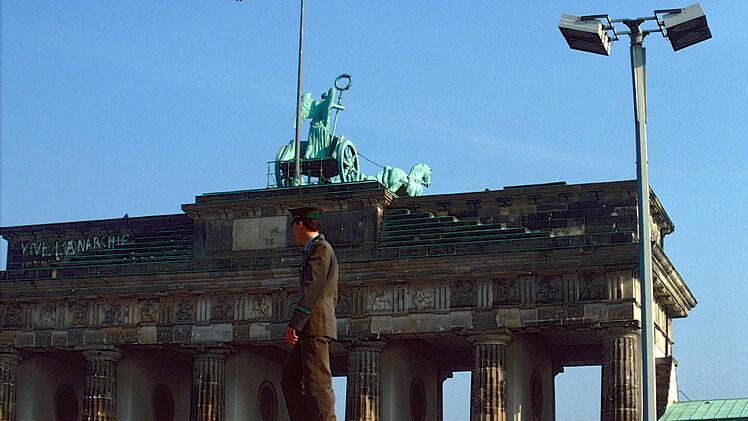 Ein DDR-Grenzer oder Volkspolizist steht vor dem Brandenburger Tor auf der Mauer, die schon sichtbar lädiert ist.