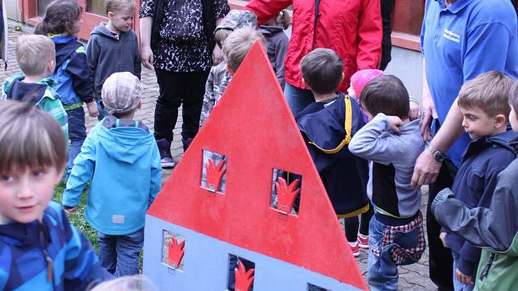 Ein Hausmodell, in dessen Fenster Flammen zu sehen sind. Willi Hussendörfer und Irmi Münch erklären den Kindern, dass sie mit einem Wasserstrahl die Flammen wegspritzen können.