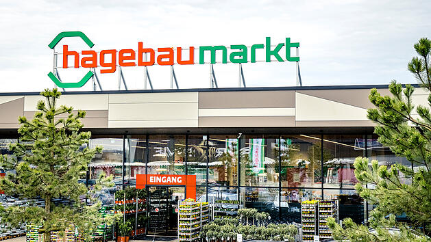 Ein Hagebaumarkt-Eingang, moderne Glasfassade, gr&uuml;ne Pflanzen und Schilder. Hell beleuchtet und von B&auml;umen umgeben.