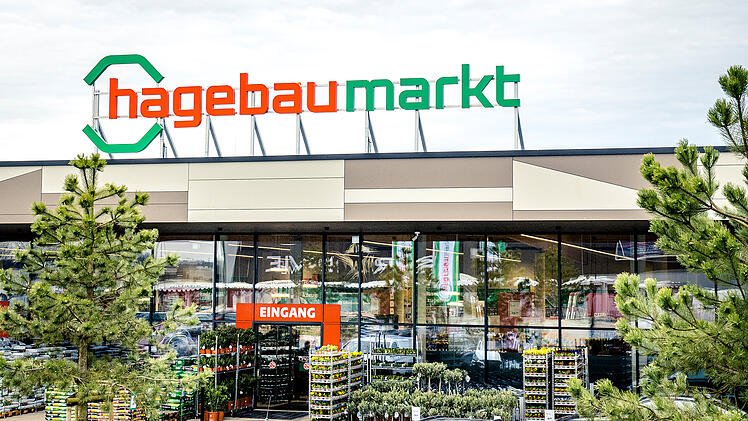 Ein Hagebaumarkt-Eingang, moderne Glasfassade, grüne Pflanzen und Schilder. Hell beleuchtet und von Bäumen umgeben.