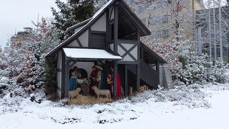 Eine verschneite Weihnachtskrippe mit traditionellen Figuren steht vor einem Fachwerkhaus, umgeben von schneebedeckten Bäumen. Im Hintergrund sieht man moderne Gebäude.