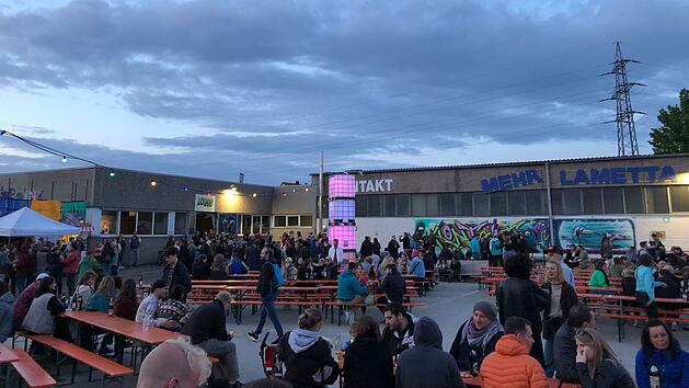 Eine Menschenmenge sitzt auf B&auml;nken bei einem Festival, umgeben von Graffiti und Lichtern. Es herrscht eine gesellige Atmosph&auml;re unter freiem Himmel.