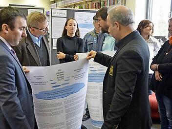 Imam Niyazi Cabbar und Ramazan Kurt halten eines der Plakate für die Ausstellung "Gebete im Dialog". Dahinter Religionslehrerin Nina Rossa, die die interreligiösen Gebete mit Schülern des Arnold-Gymnasiums erarbeitet hat. Foto: Rainer Lutz