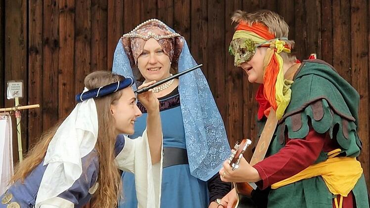 Drei Menschen in mittelalterlicher Kleidung agieren humorvoll. Einer trägt eine Gitarre, eine Frau hält einen Schnurrbart-Requisit an ihr Gesicht. Alle wirken fröhlich und engagiert.
