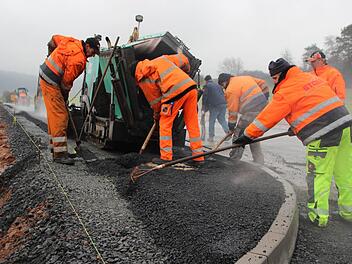 Jetzt muss es schnell gehen, ehe die Walze anrollt. Mit vereinten Kräften häufeln die Arbeiter den heißen Asphalt um einen Kanalschacht herum an. Foto: Eckehard Kiesewetter