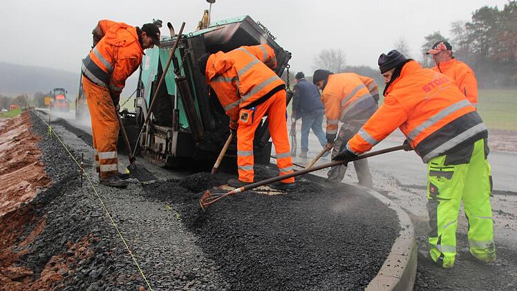 Jetzt muss es schnell gehen, ehe die Walze anrollt. Mit vereinten Kräften häufeln die Arbeiter den heißen Asphalt um einen Kanalschacht herum an. Foto: Eckehard Kiesewetter