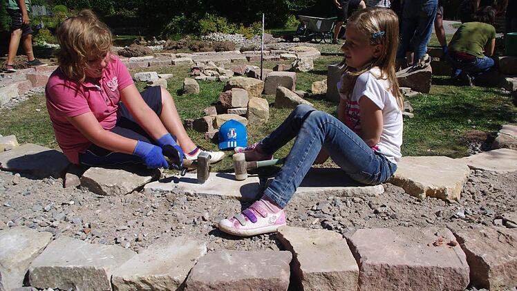 Linda Kopp und Sofia Fischer (von links) schlagen mit einem Hammer auf große Steine, damit das Fundament des Kräuter-Rondells eben wird.